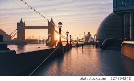 Sunrise casts warm hues over Tower Bridge as pedestrians stroll along Queen's Walk. The iconic landmark stands majestically against the brightening sky, showcasing London's beauty. 127894248