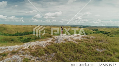 Young woman is standing on a rock admiring the breathtaking landscape of rolling green hills and cloudy sky in Sumba Island, Indonesia, embodying a sense of freedom and adventure Young woman is standing on a rock admiring the breathtaking landscape of rolling green hills and cloudy sky in Sumba Island, Indonesia, embodying a sense of freedom and adventure 127894917