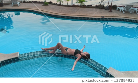 Young woman wearing bikini relaxing in infinity pool overlooking beautiful tropical beach at sunset, enjoying summer vacation in luxury resort 127894925