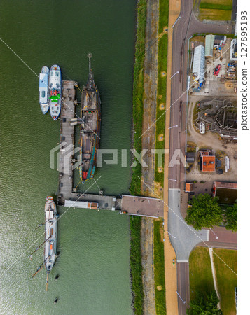 An aerial view of a coastal town with a harbor, colorful buildings and a replica ship docked along the water. Surrounding areas include parking lots and green spaces. An aerial view of a coastal town with a harbor, colorful buildings and a replica ship docked along the water. Surrounding areas include parking lots and green spaces. 127895193