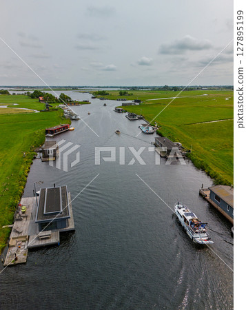 A picturesque aerial view of a Dutch windmill beside a canal with boats, surrounded by lush green fields, traditional houses, and distant waterways under a cloudy sky. 127895199
