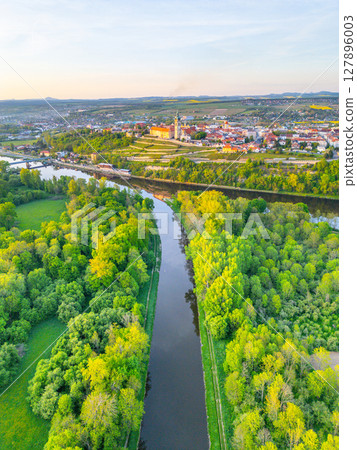 This aerial view captures Melnik City with its historic chateau perched above the Labe River. Lush greenery surrounds the riverbanks, showcasing the natural beauty of Czechia during sunset. 127896003