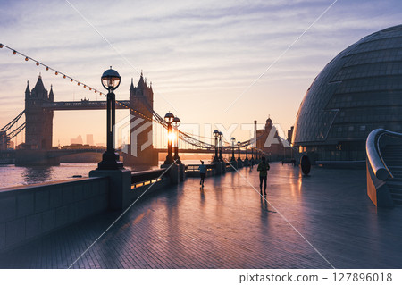 Morning sunlight casts a golden glow on Tower Bridge as people stroll along The Queen's Walk in London, enjoying the serene waterfront and iconic views of the city. 127896018