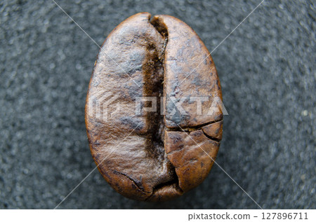 Perfectly symmetrical coffee bean in sharp focus, resting on a matte dark surface, showcasing its full shape and roast line 127896711