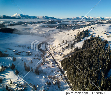 Enchanting aerial view of snow-covered village nestled among rolling hills, dense forests. Winding road cuts through scene, with morning fog drifting over valley. Majestic mountains stand in distance. Enchanting aerial view of snow-covered village nestled among rolling hills, dense forests. Winding road cuts through scene, with morning fog drifting over valley. Majestic mountains stand in distance. 127896960