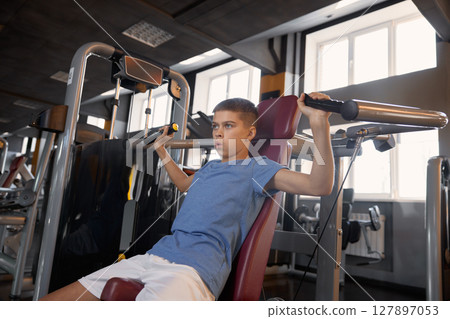 Teen boy doing sports exercise at machine for chest muscles pumping 127897053