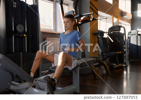 Teenage boy training press at rowing machine at modern fitness center 127897055