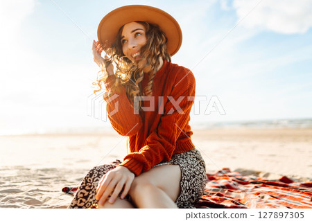 Young woman in cozy sweater, hat enjoys beautiful view of sea at beach picnic. Concept of relaxation 127897305