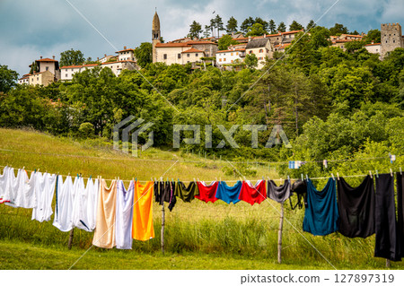 Scenic Hilltop Town Of Stanjel In Slovenia: Traditional Stone Houses And Historic Church Tower Rising Above Lush Greenery Under A Dramatic Cloudy Sky In The Karst Region 127897319