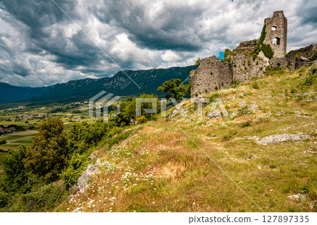 Ruins Of Grad Vipava In Slovenia: Ancient Stone Walls Covered In Ivy Standing On A Grassy Hilltop Under A Dramatic Cloud-Filled Sky In The Scenic Vipava Valley 127897335