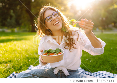 Woman enjoying a poke bowl on sunny day in a park while sitting on a picnic blanket. Concept picnic Woman enjoying a poke bowl on sunny day in a park while sitting on a picnic blanket. Concept picnic 127897434