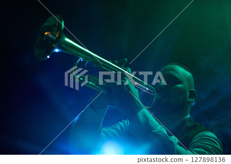 Musician playing trumpet on dimly lit stage with blue and green spotlights and atmospheric smoke. 127898136