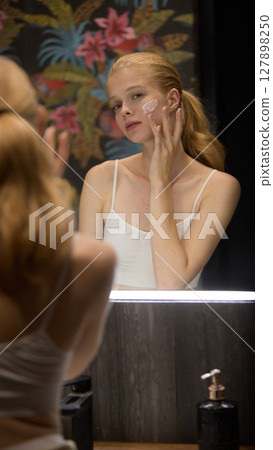 Clean-girl self-care. Woman applying facial cream in front of illuminated mirror in bathroom 127898250