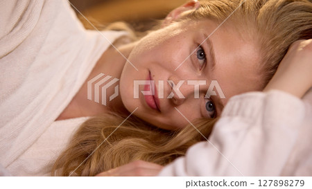 Skin-realism. Woman relaxing in cozy bed with soft white robe and serene expression 127898279