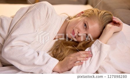 Woman relaxing comfortably in plush white robe on soft bed at home interior Woman relaxing comfortably in plush white robe on soft bed at home interior 127898280