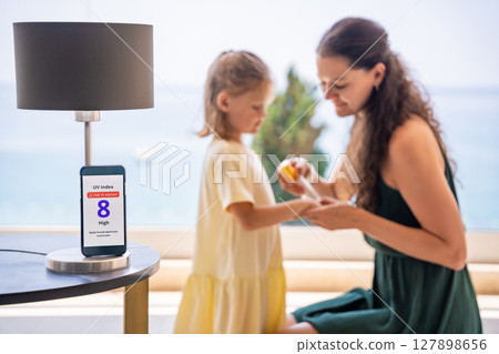 Smartphone with high UV alert in focus, while mother applies sunscreen to daughter in background. Real-life response to sun safety warning Smartphone with high UV alert in focus, while mother applies sunscreen to daughter in background. Real-life response to sun safety warning 127898656