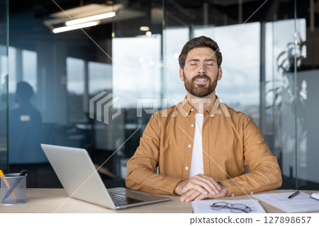 A man in an office setting with his eyes closed, looking relaxed, with a laptop and office supplies on the desk. 127898657