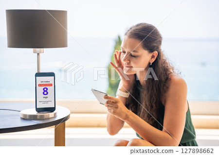 Smartphone with high UV alert in focus, while woman applies sunscreen to her face in mirror. Conscious skincare routine inspired by sun safety warning. Smartphone with high UV alert in focus, while woman applies sunscreen to her face in mirror. Conscious skincare routine inspired by sun safety warning. 127898662