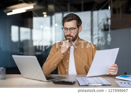 A thoughtful businessman in glasses reviews documents at his desk, with a laptop and calculator nearby. The office setting includes a blurred background. A thoughtful businessman in glasses reviews documents at his desk, with a laptop and calculator nearby. The office setting includes a blurred background. 127898667
