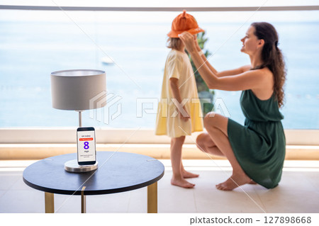 Mother puts sun hat on daughter as smartphone displays high UV alert. Practical response to sun safety warning for child protection 127898668