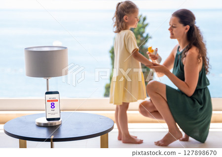 Smartphone with high UV alert in focus, while mother applies sunscreen to daughter in background. Real-life response to sun safety warning 127898670