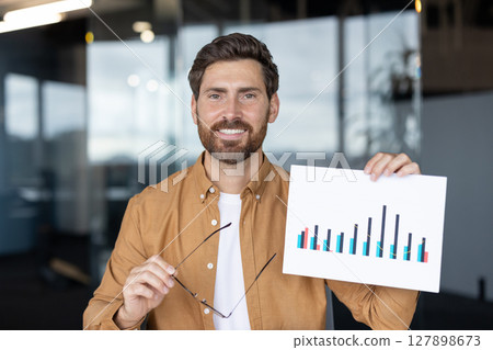 A man smiles during an online presentation, holding glasses and a chart with graphs, representing data analysis in a modern office environment 127898673