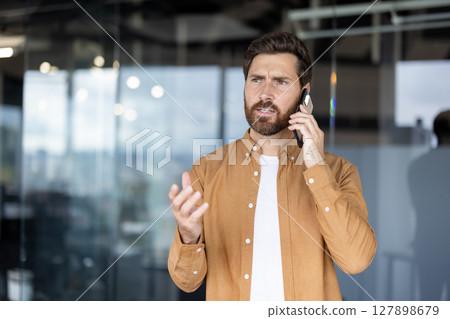 A concerned businessman communicates on his smartphone, gesturing animatedly while looking upwards in an office setting. 127898679
