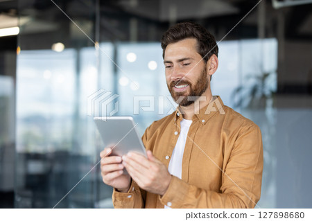 A smiling businessman holds a tablet in a modern office setting, demonstrating engagement and technology use. 127898680