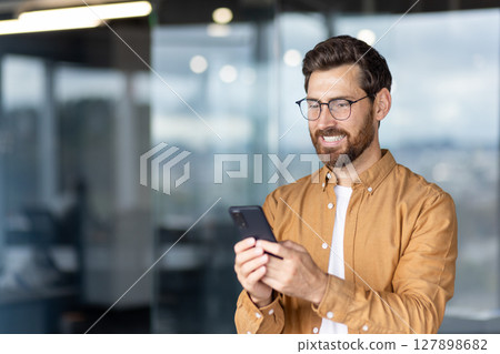 A smiling man in glasses looks at his phone while in an office setting with a blurred background. 127898682