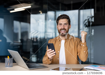 A happy businessman celebrating success while holding a smartphone in an office environment. 127898696