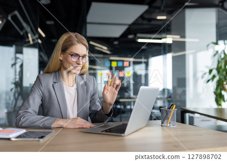 Online video conference, successful businesswoman boss inside office at workplace using laptop for video call. Woman in business suit waving hand greeting colleagues and partners. Business meeting. 127898702
