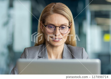 Confident businesswoman wearing glasses focused on working with laptop in modern office. Demonstrates professionalism and concentration while engaging with digital work environment 127898714
