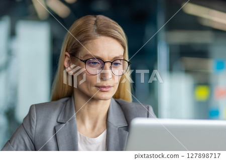 Confident businesswoman wearing glasses concentrating on work at laptop in modern office space. Professional attire reflects office environment, representing focus and determination in work setting. Confident businesswoman wearing glasses concentrating on work at laptop in modern office space. Professional attire reflects office environment, representing focus and determination in work setting. 127898717