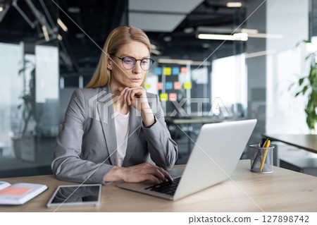 Mature woman in a professional setting focused on her laptop while seated at a desk. The image suggests concentration, productivity, and a corporate atmosphere. 127898742