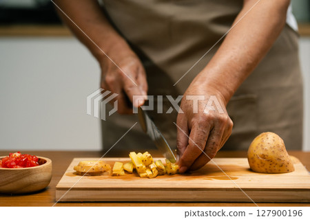 Close-up of Asian man's hands slicing potatoes on a wooden cutting board in the kitchen, preparing for cooking. 127900196