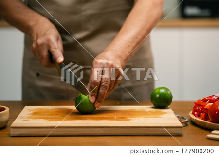 Close-up of man's hand cutting lemon on wooden cutting board in kitchen, preparing for cooking. 127900200