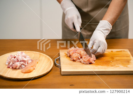 Close-up of Asian man's hands cutting chicken on a wooden cutting board in the kitchen, preparing for cooking. Close-up of Asian man's hands cutting chicken on a wooden cutting board in the kitchen, preparing for cooking. 127900214