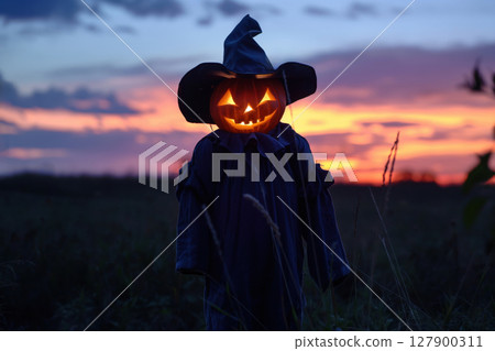 Scarecrow with a pumpkin head and a glowing candle standing in a field at sunset 127900311