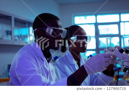 Two scientists wearing lab coats and safety glasses working together in a laboratory, carefully examining a test tube 127900728
