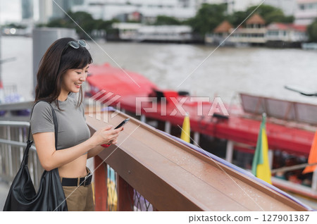 Happy young southeast asian woman teenager blogger explorer using smartphone searching tourism guide for local cultural experience. Asia world travel concept in Bangkok by Chao Phraya River 127901387