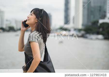 Happy solo Southeast Asian woman teenager travel blogger uses smartphone. An explorer enjoying local cultural experience Asia waiting for friend or express boat by Chao Phraya River Bangkok Happy solo Southeast Asian woman teenager travel blogger uses smartphone. An explorer enjoying local cultural experience Asia waiting for friend or express boat by Chao Phraya River Bangkok 127901410