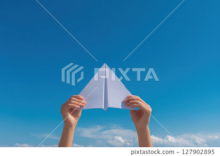 Hands Holding a Paper Plane Against a Clear Blue Sky in Daylight with Soft Clouds 127902895