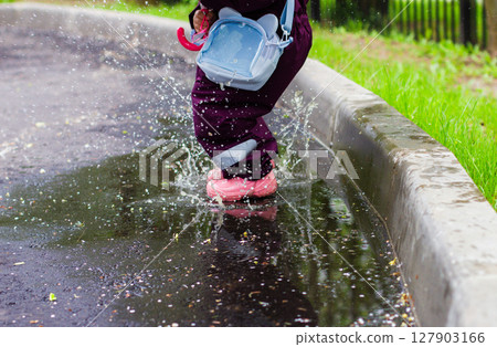 The child jumps through the puddles. The child in a bright cloak and colorful rubber boots joyfully jumps and splashes in a puddle next to the sidewalk, holding a small pink umbrella on a rainy day. 127903166