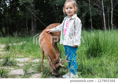 Nestled in a tranquil forest, a young girl gently petting a roe deer captures a heartwarming bond between humans and wildlife, highlighting moments of kindness and connection in nature. 127903168