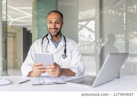 Successful experienced doctor sitting at desk inside the clinic office and using a tablet computer. Man in a white medical coat, using an application for online communication and consulting patients. 127904687