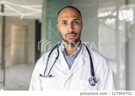 A close-up portrait of a professional, confident Black doctor in a medical setting, wearing a white coat and stethoscope. 127904702