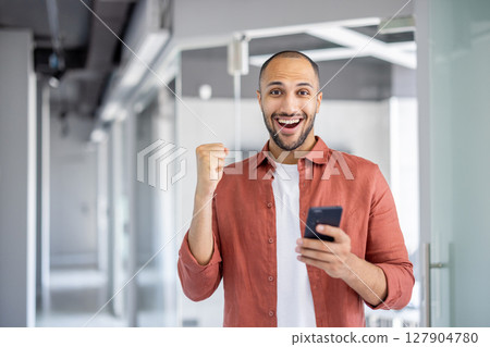 A man celebrates success, holding his phone with an excited expression in a modern office setting. A man celebrates success, holding his phone with an excited expression in a modern office setting. 127904780