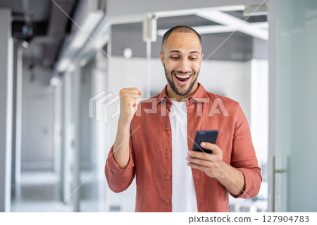 A joyful man in a shirt looks at his phone with an elated expression, celebrating a win or positive news indoors. A joyful man in a shirt looks at his phone with an elated expression, celebrating a win or positive news indoors. 127904783