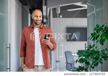 Successful businessman walking through the office with a phone in his hands, the man is typing a text message and smiling, browsing social networks and internet pages at his workplace. Successful businessman walking through the office with a phone in his hands, the man is typing a text message and smiling, browsing social networks and internet pages at his workplace. 127904785