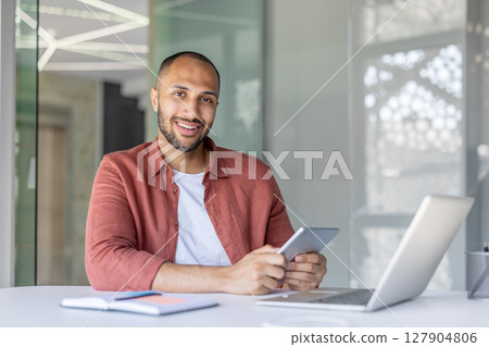 Portrait of a young successful man office worker programmer developer, looking at the camera smiling, with a tablet computer in his hands. Sitting at a desk with a laptop. Portrait of a young successful man office worker programmer developer, looking at the camera smiling, with a tablet computer in his hands. Sitting at a desk with a laptop. 127904806